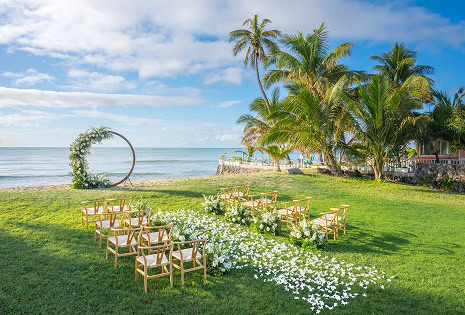 青空と海、ヤシの木を背景にした、白い花びらのバージンロードと丸いフラワーアーチが設置された開放的なビーチサイドの屋外結婚式会場